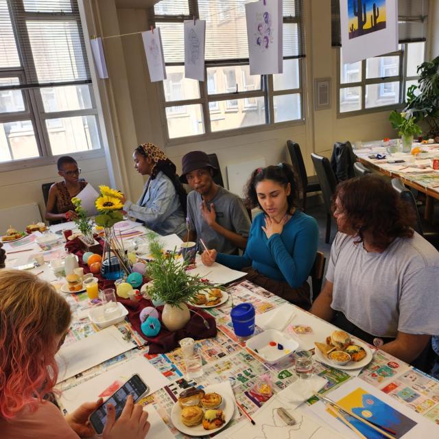 A group of individuals sitting around a table covered with newspapers, art supplies, and food items like fruits and sandwiches. A vase with sunflowers is in the center. Some people are crafting, while others are eating or conversing. The room has posters on the walls and large windows letting in natural light.