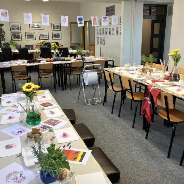 An art workshop with tables arranged in a U-shape, covered with paintbrushes, palettes, and watercolor papers. The room is colorful, with paintings on the walls and easels. A green vase with yellow flowers is in the center.