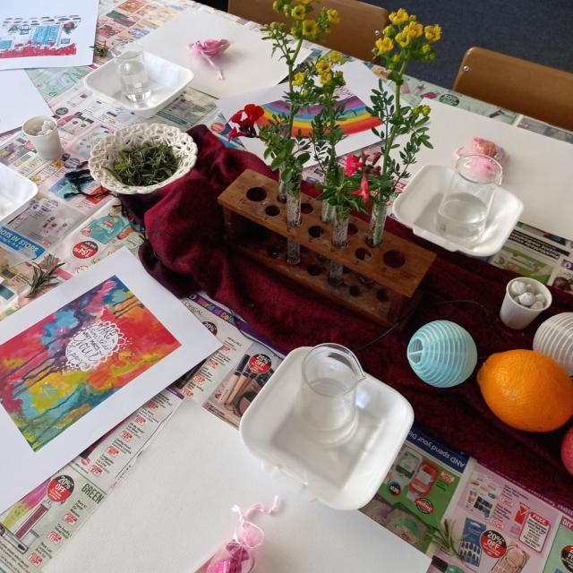 A table set up for a craft or educational activity with a wooden block holding colorful flowers, teacups, a teapot, greenery, paint supplies, printed pictures, oranges, and a blue paper lantern on a tablecloth with text and images.