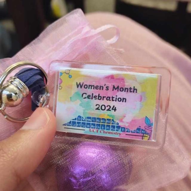 A hand holding a small, rectangular badge with a keychain. The badge reads ‘Women’s Month Celebration 2024’ with colorful abstract designs and a chemistry-related logo. The badge is on a pink fabric surface.