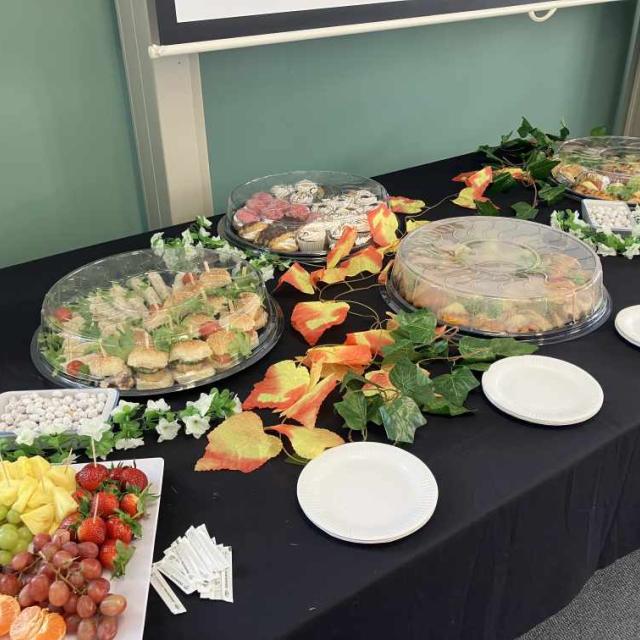 A buffet table set up for an event with platters of sandwiches, wraps, fruits, and vegetables. Decorative foliage with autumn leaves accents the presentation. A chalkboard and a bottle of orange juice are in the background.