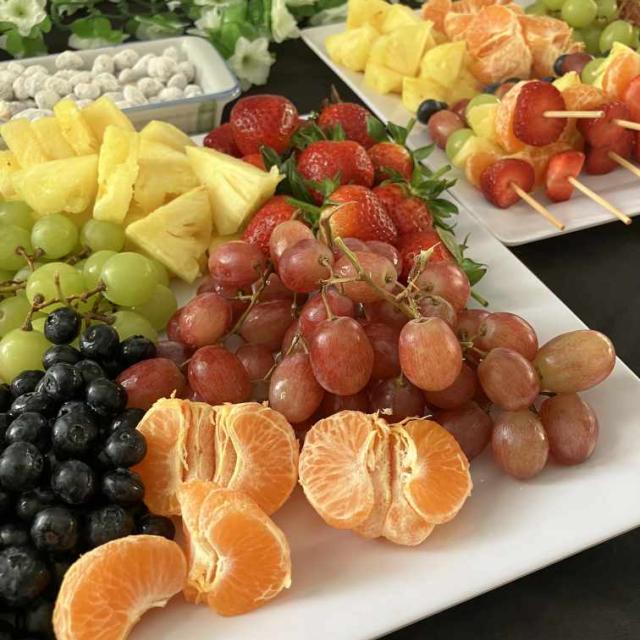 A white platter filled with a variety of fresh fruits, including red and green grapes, pineapple slices, strawberries, blackberries, and mandarin orange segments. In the background, there are fruit skewers and a bowl of nuts or small snacks on a dark surface with white flowers in the upper left corner.