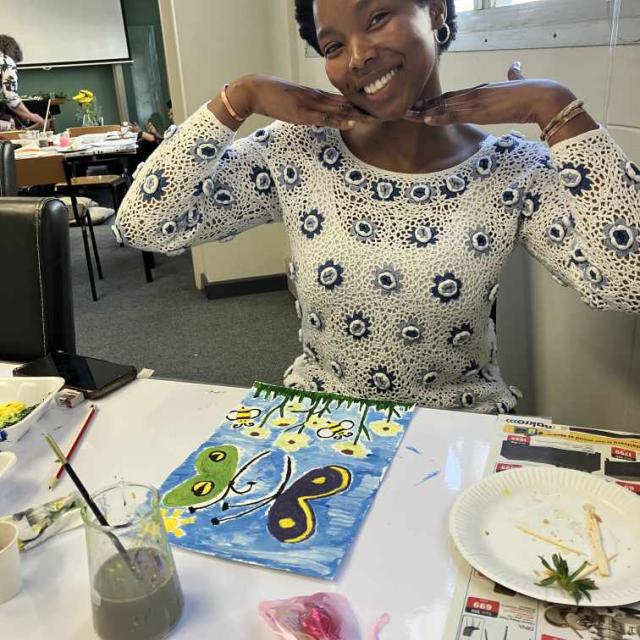 An individual is sitting at a table with a painting of a blue butterfly with yellow and green details on a background of green grass and blue sky, with yellow flowers at the bottom. The table has paintbrushes, a jar of water, and various colors of paint, suggesting an art class or workshop setting.