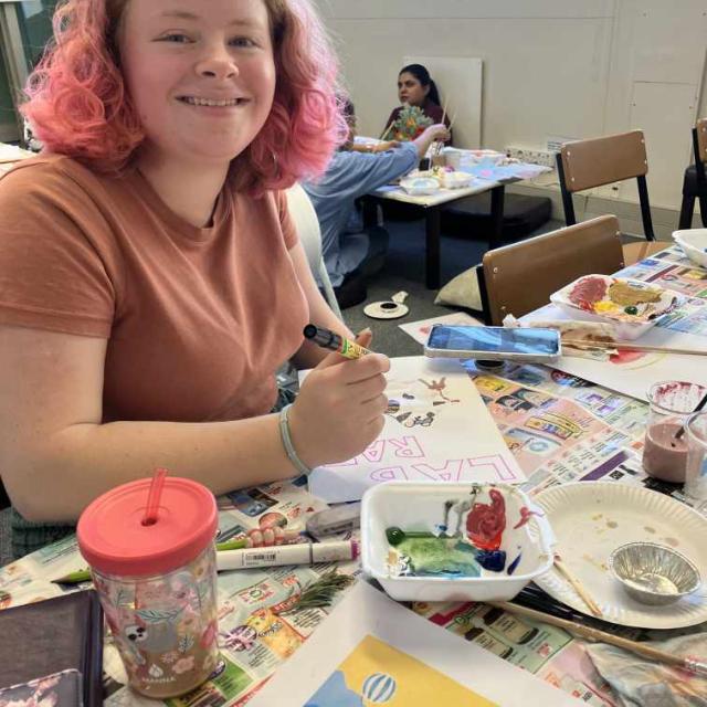 An individual is sitting at a table covered with newspapers, engaged in painting. Various art supplies, including paint palettes and brushes, are scattered around. In front of the individual is a colorful artwork featuring blue and yellow shapes on paper, suggesting an art class or workshop setting.
