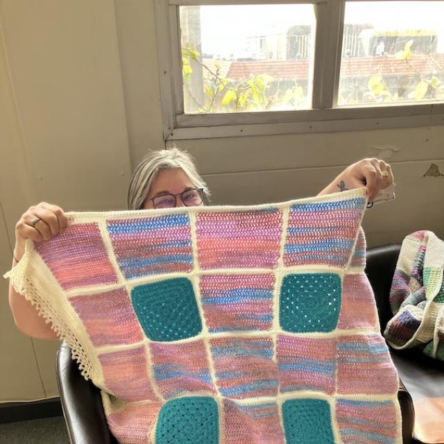 woman with glasses holding up a pink and blue knitted blanket