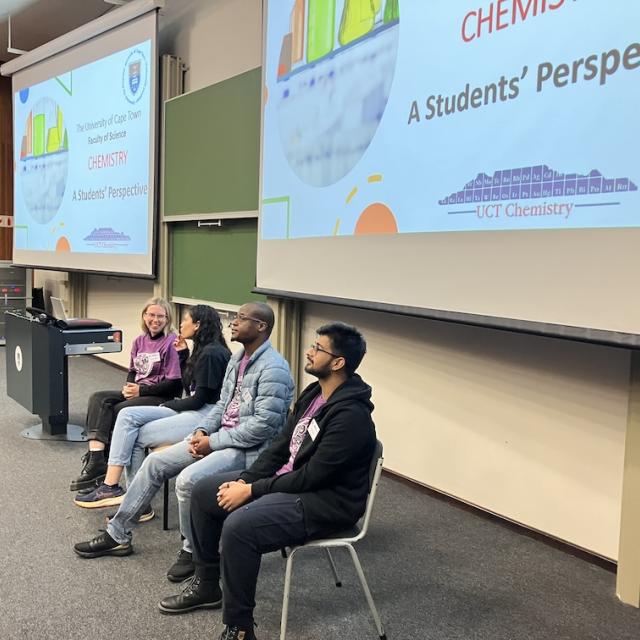Four chemistry department postgraduate students sit on chairs in front of lecture theatre boards