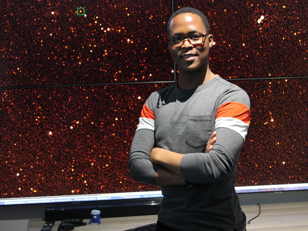 Mr Reikantseone Diretse in front of the MeerKAT observation of GRB190114C (encircled) in the IDIA Visualisation Lab. (credit: Siphelo Funani)