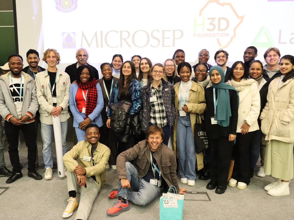 A group of people standing together and smiling. The background shows a white screen with the logos ‘EMICROSEP’ and ‘H3D FOUNDATION’.