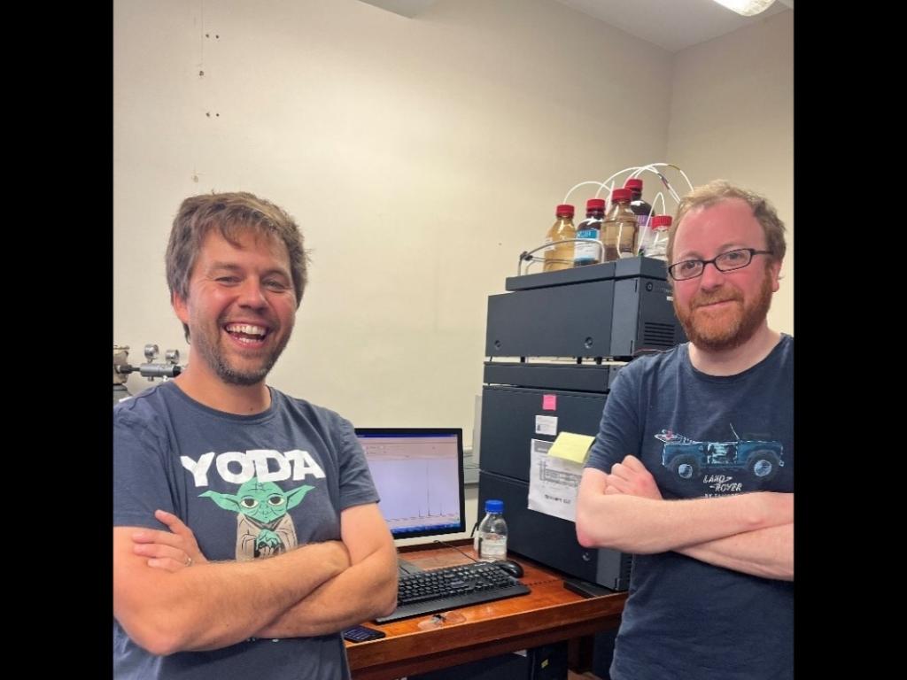 Picture of two smiling men standing in front of a mass spectrometer.