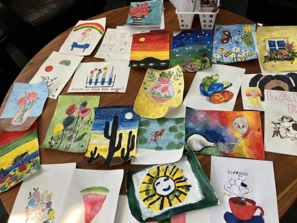 A collection of colorful children’s artwork displayed on a wooden surface, featuring various subjects like landscapes, animals, plants, and abstract designs. Some pieces include motivational text such as ‘BLOOM IN YOUR OWN TIME’ and ‘BE HAPPY BE BRIGHT BE YOU.’