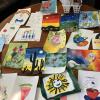 A collection of colorful children’s artwork displayed on a wooden surface, featuring various subjects like landscapes, animals, plants, and abstract designs. Some pieces include motivational text such as ‘BLOOM IN YOUR OWN TIME’ and ‘BE HAPPY BE BRIGHT BE YOU.’